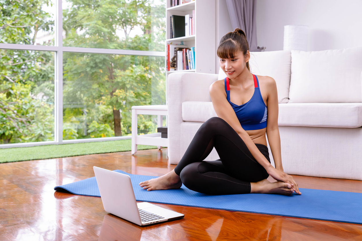 Lady stretching on a yoga mat whilst watching workout videos on her laptop.