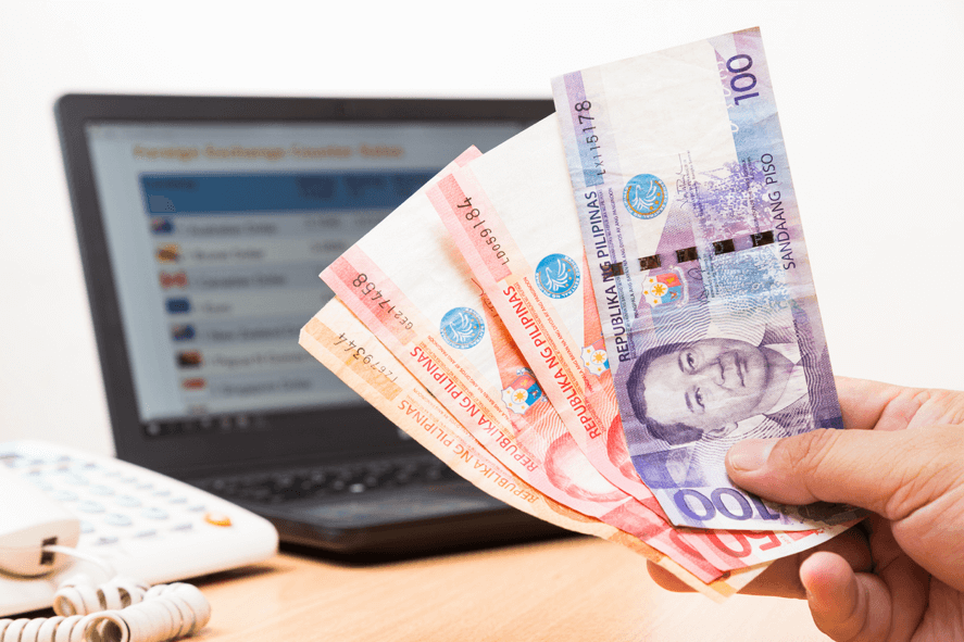 Send Money to the Philippines Hand holding Philippine peso bills with laptop and telephone in the background.