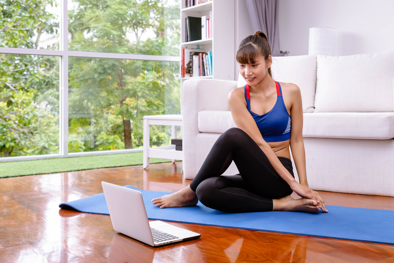 Lady stretching on a yoga mat whilst watching workout videos on her laptop.