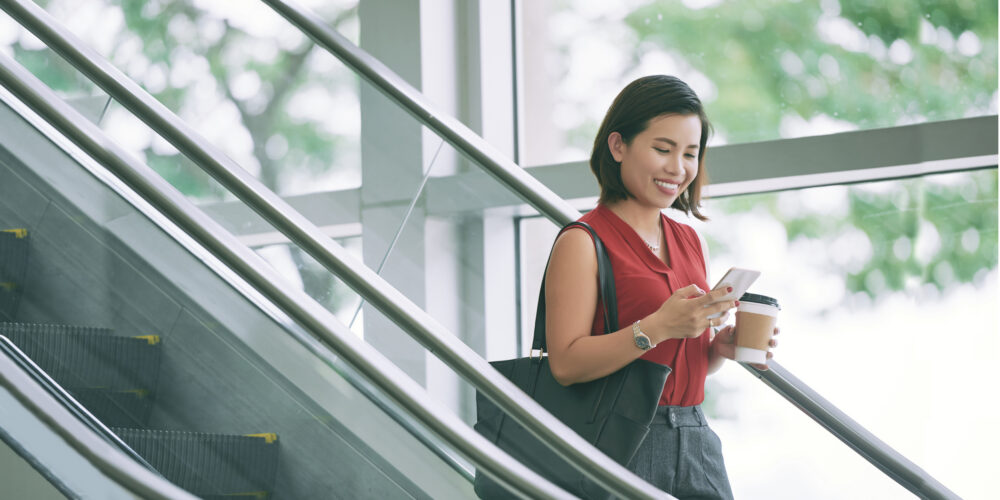 Business woman checking phone Pretty smiling woman with coffee cup reading text messages when standing on escalator