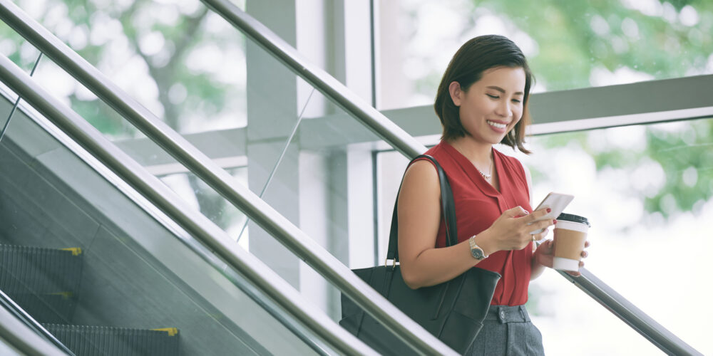 Business woman checking phone Pretty smiling woman with coffee cup reading text messages when standing on escalator